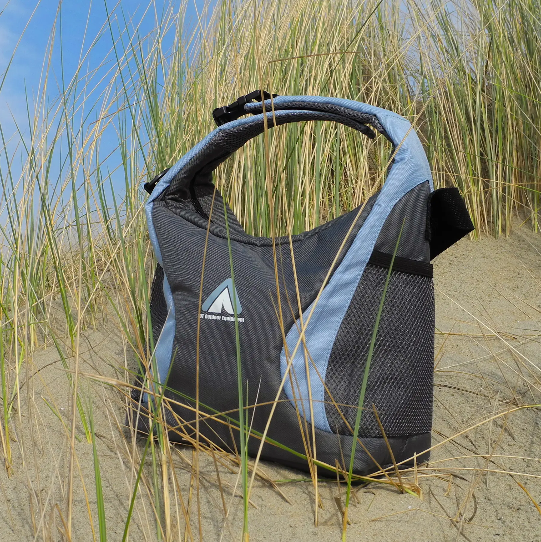 Ein schwarze und blaue Outdoor-Tasche steht auf einem Sandstrand zwischen Gräsern vor einer blauen Himmelshintergrund.