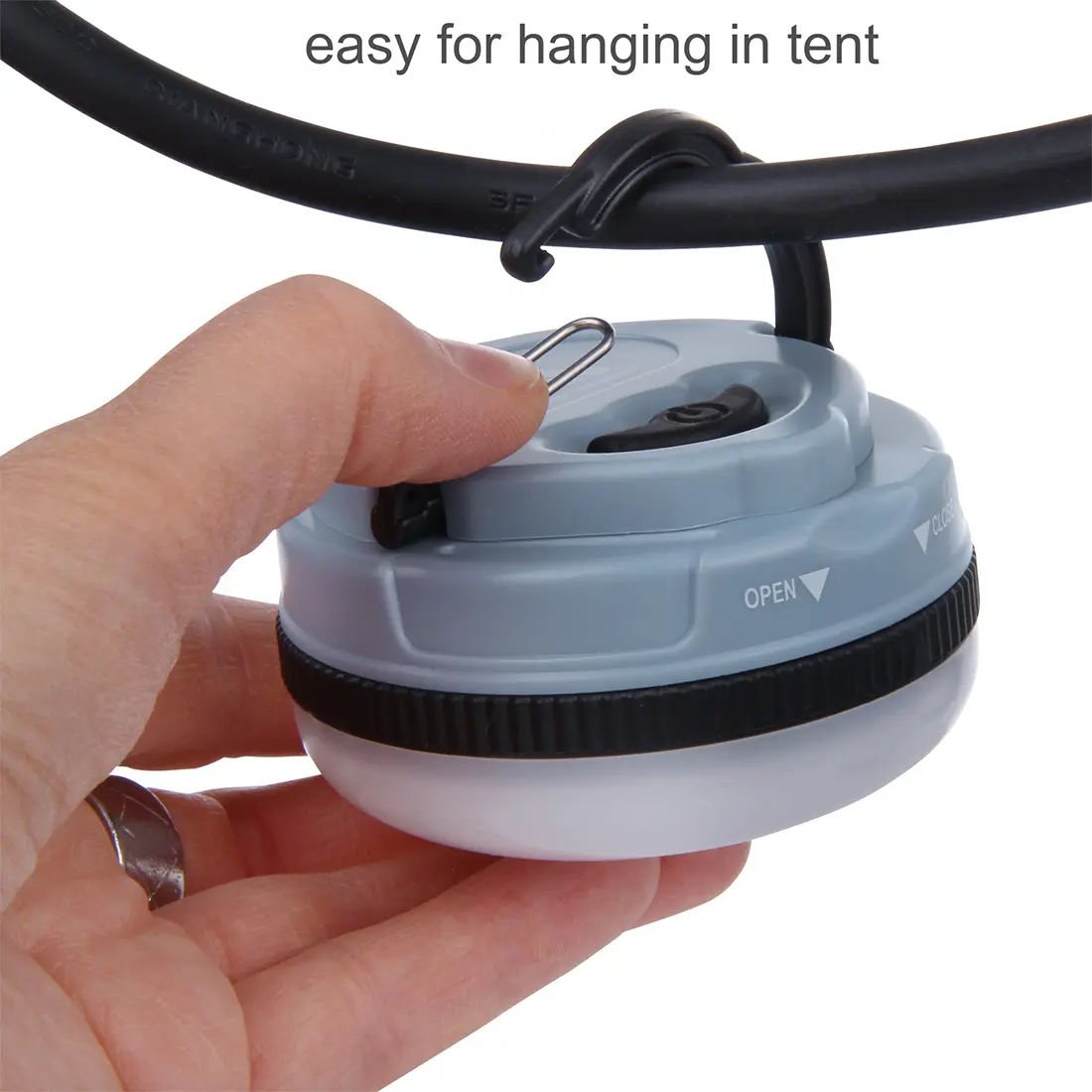 Die Abbildung zeigt eine Hand, die einen kleinen, runden, grauen und schwarzen Aufhängeschalter mit einem Metallhaken an einem Hängeventil befestigt, über dem die Text "easy for hanging in tent" steht.