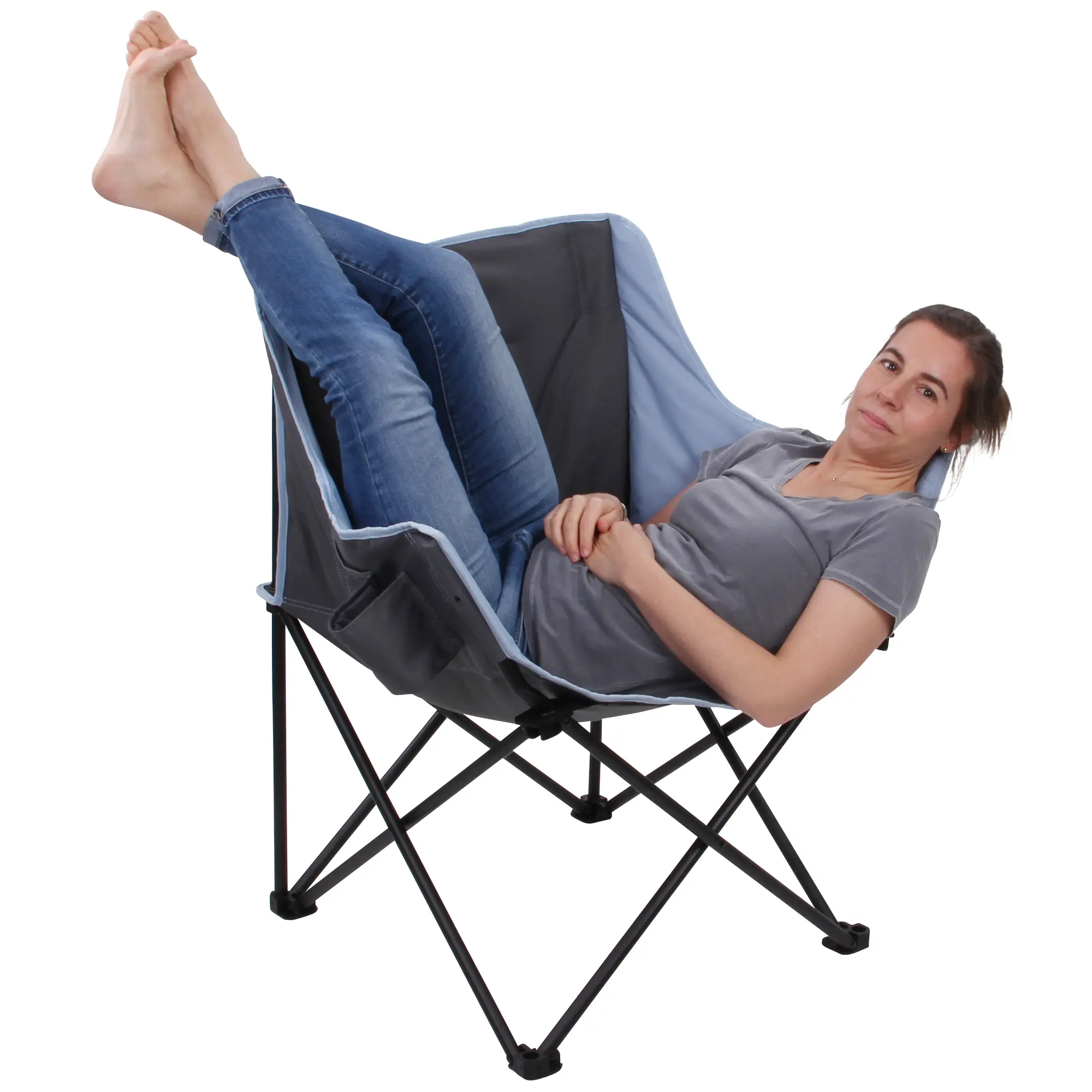 Eine Frau liegt entspannt in einem zusammengefalteten Campingstuhl, trägt eine grau T-Shirt und blaue Jeans, und schaut lächelnd in die Kamera.