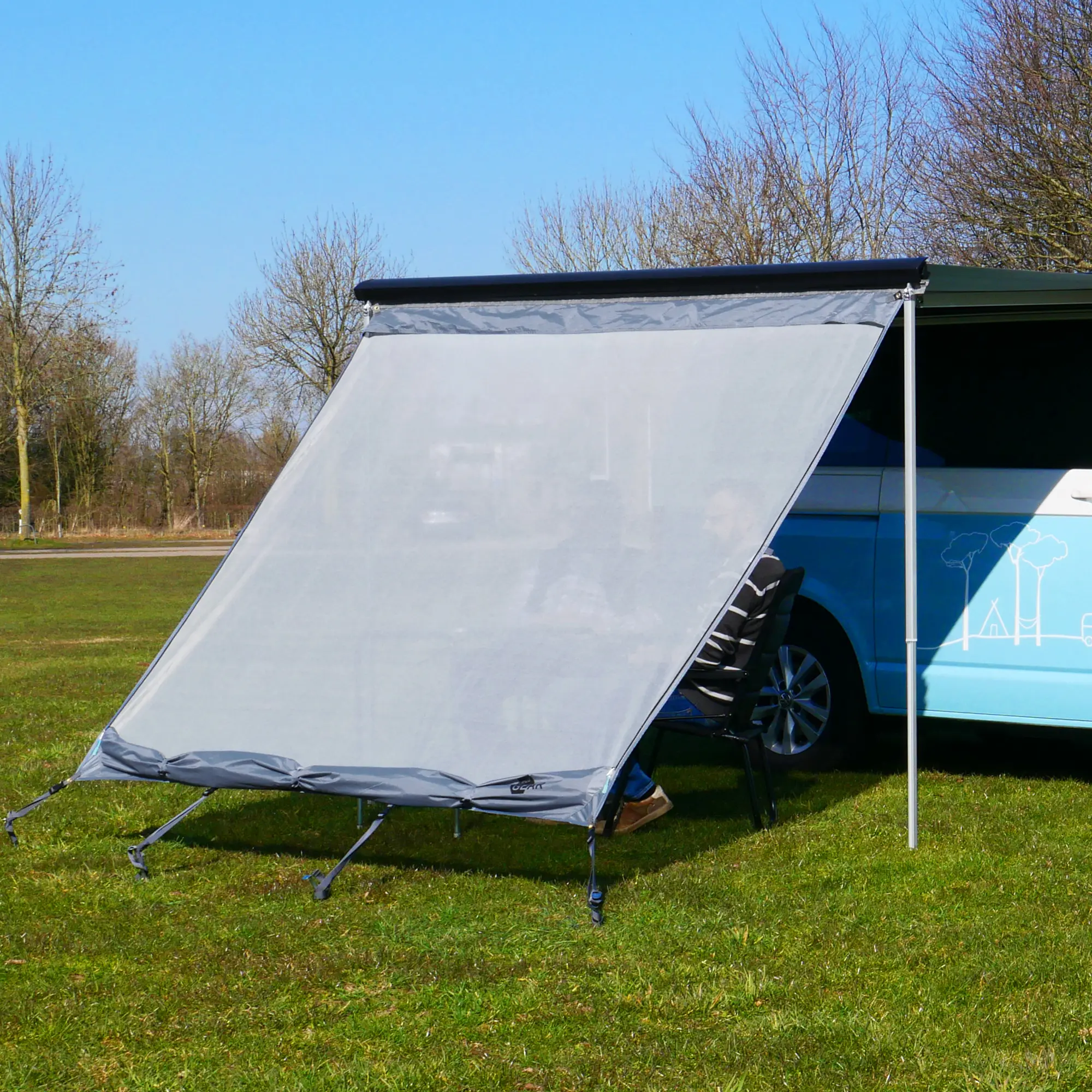 your GEAR Campingbus Sun + Rain Blocker 2,4 x 2 m - Vorderwand für Bulli / Bus Markise mit Abdeckung Ein blau-weißer Camper steht auf einer Wiese mit einer ausgerollten Markise, die durch Stangen gestützt wird, während ein Mann auf einem schwarzen Stuhl im Schatten sitzt, umgeben von kahlen Bäumen unter einem klaren blauen Himmel.